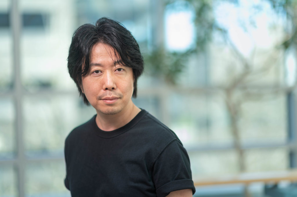 Man standing in front of blurred office background. He has slightly long dark hair and is wearing a short sleeved black shirt. He is standing with his right shoulder slightly pointing away from the camera field of view.