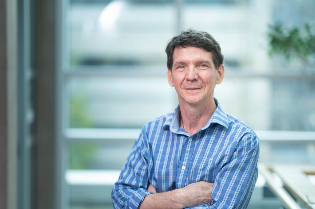 Man standing in front of blurred office background, there is a blurred tree in the top right hand corner. He has short dark hair and is wearing a blue striped shirt, his arms are crossed.