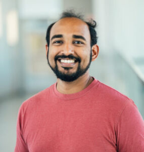 Headshot of Manu Madhav. He is wearing a red long sleeved sweater.